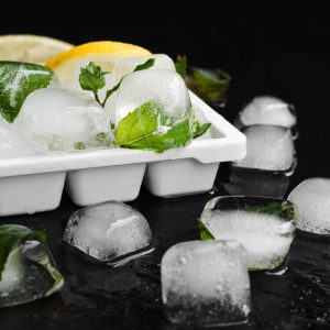 An image of an Ice tray filled with ice cubes on a black marble table with slices of lemon and mint